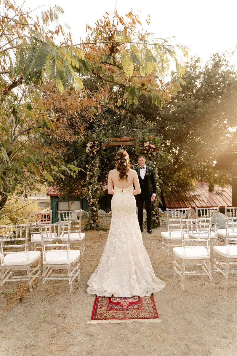 A bride walking the vintage rug aisle toward her groom in backlit afternoon sun